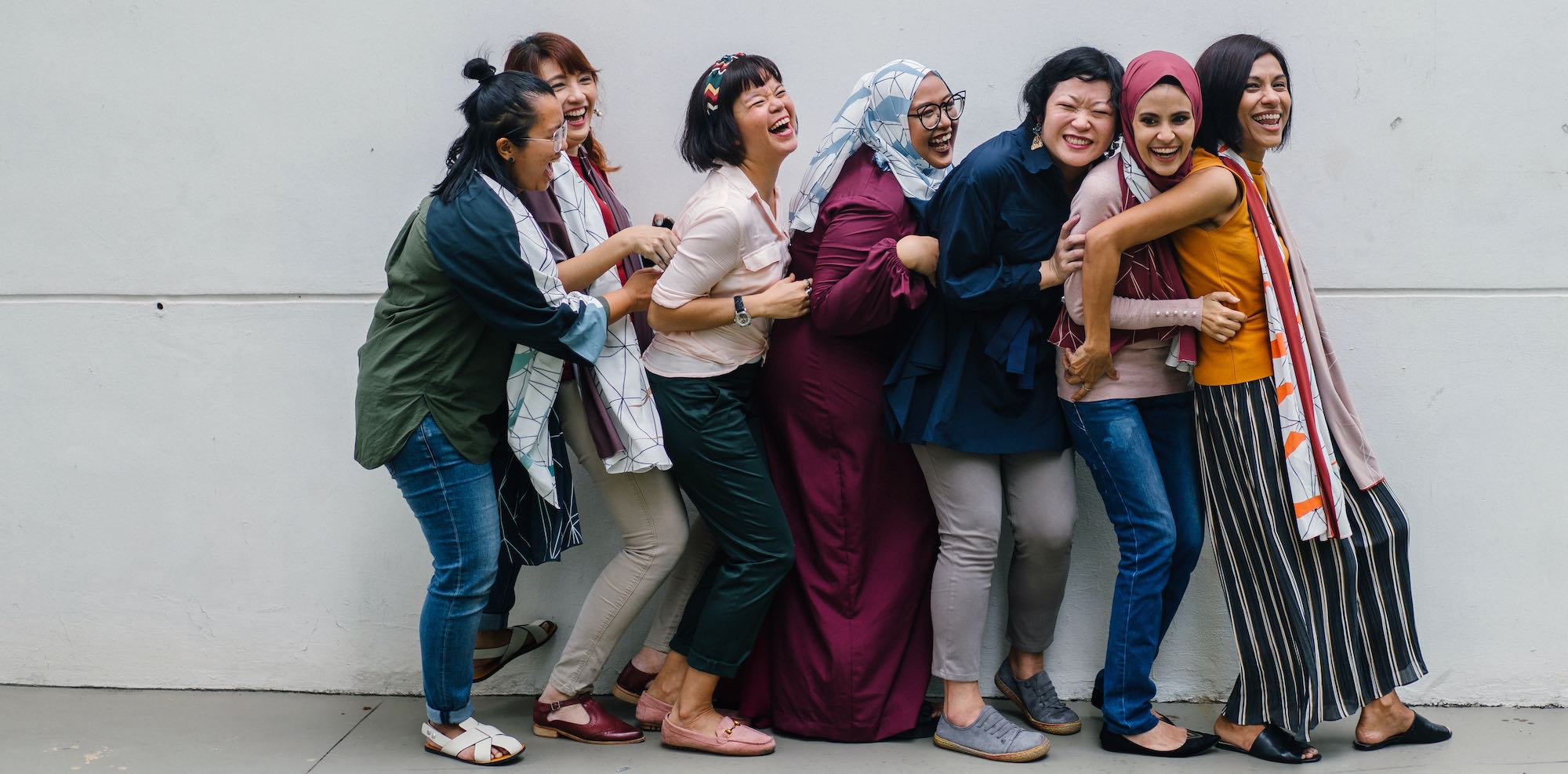 seven women standing against a white wall laughing