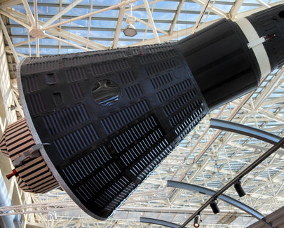 A black and silver Mercury space capsule displayed indoors at a museum, suspended from the ceiling with a glass roof and metal framework in the background
