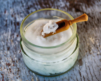 A glass jar filled with solidified coconut oil, with a small wooden spoon scooping some out, placed on a rustic wooden surface.
