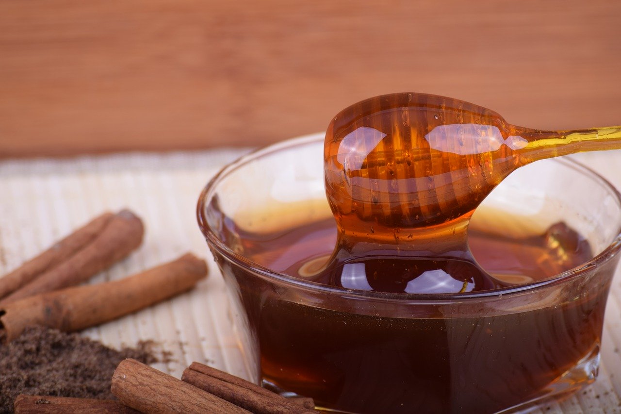 A glass bowl of amber honey with a wooden honey dipper and cinnamon sticks on a wooden surface